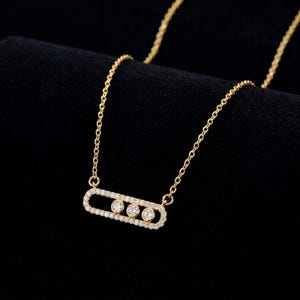 May include: A gold necklace featuring a horizontal, oval-shaped pendant frame encrusted with small clear stones. Inside the oval frame, three larger, round, clear stones are set in a row. The pendant is attached to a delicate gold chain.