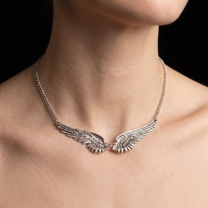 Op de afbeelding: Een zilverkleurige metalen ketting met een centrale hanger in de vorm van gespreide vleugels. De vleugels zijn verbonden met een fijne zilverkleurige ketting die om de nek rust. Het metaal heeft een textuur met gedetailleerde veerpatronen.