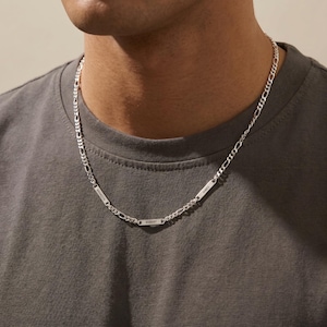 May include: A silver Figaro chain necklace with three rectangular nameplates. The nameplates are engraved with the names "BRANDON", "ROBERT", and "JAMES". The necklace is worn around the neck.