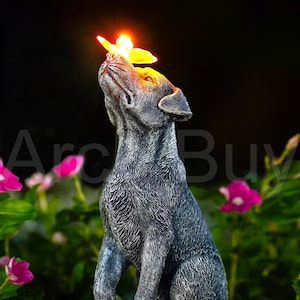 May include: A gray dog statue with a butterfly on its nose, illuminated with a warm, yellow light. The dog is sitting, looking upwards. Pink flowers and green foliage are in the background.