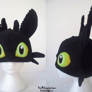 Puede incluir: Gorro negro que se asemeja al personaje Desdentado de la película "Cómo entrenar a tu dragón". El gorro presenta ojos grandes, verdes y amarillos, y detalles distintivos de orejas y aletas. El gorro se muestra en una cabeza de maniquí blanca.