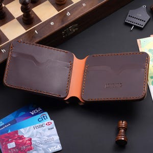 May include: Open brown leather wallet with an orange interior and visible stitching. The wallet features multiple card slots and the brand name "LOXBURG" is embossed. Credit cards are visible in the background.