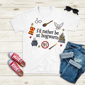 Peut inclure: Un t-shirt blanc avec le texte noir "i'd rather be at hogwarts" entouré d'illustrations colorées comprenant une écharpe rayée, des lunettes rondes, un balai, une chouette blanche, un château, un chaudron, une baguette et un signe "9 3/4".
