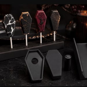 May include: Five coffin-shaped cake pops on white sticks stand in a black rectangular holder. The pops feature purple, white-marbled, gold-veined, dark red, and gold-speckled black finishes. Nearby are black silicone coffin molds and a stand.