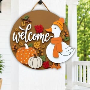 Puede incluir: Un letrero de pared redondo de madera con fondo marrón que presenta el texto en cursiva blanca welcome. El letrero incluye un ganso blanco con gorro y bufanda naranja, una calabaza naranja con lunares, una calabaza blanca y hojas de otoño.