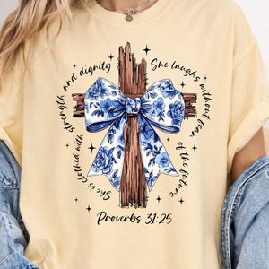 May include: A light yellow t-shirt featuring a wooden cross with a blue floral bow. The shirt has the text "She is clothed with strength and dignity" and "Proverbs 31:25" printed on it.