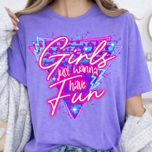 May include: A lilac-colored t-shirt with the text "Girls just wanna have fun" in a neon pink and blue design. The graphic is set within a geometric shape with lightning bolt accents, creating a retro aesthetic.