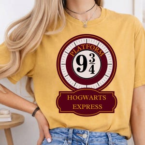 May include: A mustard yellow t-shirt featuring a circular graphic with the text Platform 9 3/4 and a rectangular banner below with the text HOGWARTS EXPRESS in gold lettering. The design uses a maroon and cream colour palette on the shirt fabric.