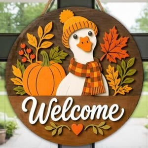 May include: A round wooden door sign featuring a white goose wearing an orange knit hat and a plaid scarf. The design includes a large orange pumpkin, autumn leaves, and red berries. The word Welcome is written in white script at the bottom.