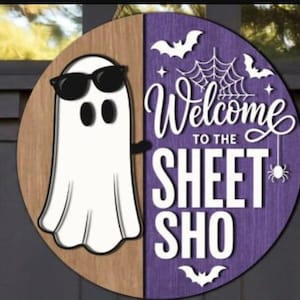 May include: A round wooden door sign split into a natural wood side and a purple side. A white ghost wearing black sunglasses is on the left. The right side features white text reading Welcome TO THE SHEET SHO, with white spiderweb, bat, and spider icons.