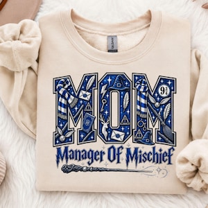 May include: A beige crewneck sweatshirt featuring the word MOM in large blue block letters filled with magical icons like a scarf, a broomstick, a wand, and a hat. Below the word MOM, the text reads Manager Of Mischief in blue font.