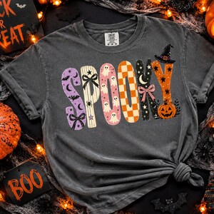 May include: A charcoal grey short-sleeve t-shirt featuring the word SPOOKY in large, patterned letters. The S is purple with bats, the P is striped with bows, the O is pink with ghosts, the O is orange checkered, the K is black with stars, and the Y is orange with a pumpkin and witch hat.