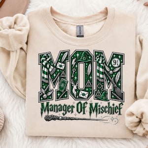 Peut inclure: Un sweat-shirt beige à col rond avec le mot MOM en grandes lettres vertes remplies d'icônes magiques comme des écharpes, des baguettes et des chapeaux. En dessous, le texte indique Manager Of Mischief en vert avec une baguette.