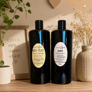 May include: Two black bottles of cleaning products with labels that read "Mop Soap" and "Gain Laundry Detergent". The "Mop Soap" bottle is 12 fl oz. The bottles are on a wooden shelf with a framed sign that says "Clean Home Happy Heart".