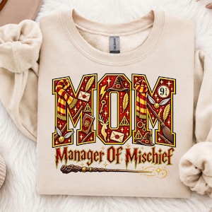 Peut inclure: Un sweat-shirt beige à col rond arborant le mot MOM en grandes lettres rouges remplies d'icônes magiques comme une écharpe rayée, un chapeau de sorcier, un balai et un vif d'or. En dessous, le texte Manager Of Mischief avec une baguette.