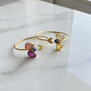Peut inclure: Deux bracelets dorés ornés de pierres précieuses. Chaque bracelet présente un motif de croissant de lune et est orné de pierres précieuses colorées, notamment des pierres roses, violettes et jaunes, et d'un petit charme en forme d'œil bleu. Les bracelets sont présentés sur une surface en marbre blanc.