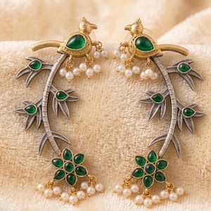 May include: A pair of ornate earrings featuring a bird design with green gemstone eyes and accents. The earrings have a silver and gold finish, with a floral and leaf design, and small pearl accents. The earrings are set against a cream-colored background.
