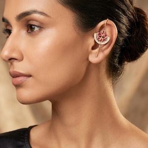 May include: A gold ear cuff featuring a fan-shaped design with red gemstones and small white stones. The ear cuff is worn on the ear and has a long, thin gold bar that curves around the ear.