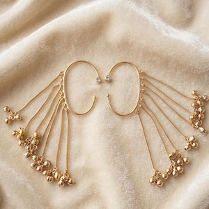 May include: A pair of gold-toned ear cuffs featuring an oval-shaped frame with a single clear stone at the top. Multiple thin gold chains hang from the frame, each ending in a small gold-toned bell charm that creates a layered, dangling effect.
