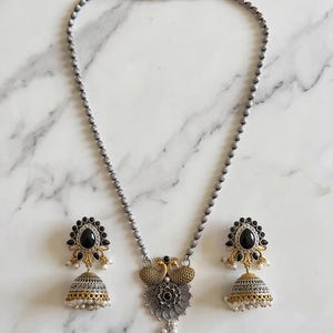 May include: A silver-toned beaded necklace featuring a central pendant with two gold-toned peacocks above a circular floral motif, accompanied by two matching jhumka earrings with black teardrop stones and small white bead accents.