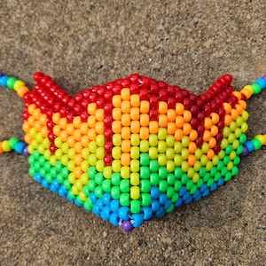 May include: A rainbow-colored beaded face mask. The mask is made of small, colorful beads arranged in a gradient pattern, transitioning from red at the top to blue at the bottom. The ear loops are also made of rainbow-colored beads.