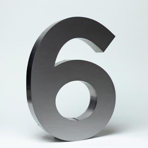 Könnte beinhalten: Eine große, metallische Zahl sechs in gebürsteter Stahlausführung. Die Ziffer ist dreidimensional und steht aufrecht vor einem schlichten weißen Hintergrund. Die Zahl hat ein modernes, minimalistisches Design.