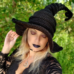 Op de afbeelding: Een zwarte gehaakte heksenhoed met een hoge, gekrulde rand. De hoed wordt gedragen door een persoon met lang blond haar en donkere make-up. De persoon draagt een zwart kanten topje en een zilveren ketting met een halve maan hanger.
