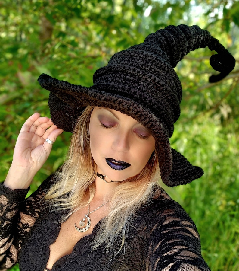 Op de afbeelding: Een zwarte gehaakte heksenhoed met een hoge, gekrulde rand. De hoed wordt gedragen door een persoon met lang blond haar en donkere make-up. De persoon draagt een zwart kanten topje en een zilveren ketting met een halve maan hanger.