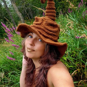 May include: A brown crochet witch hat with a wide brim and a curly, pointed top. The hat is made of a textured yarn and is worn outdoors in a natural setting with green foliage and purple flowers.