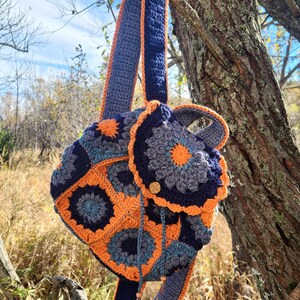 May include: A handmade crochet backpack in shades of navy blue, orange, and gray, hanging from a tree branch. The bag features a floral pattern and a flap closure with a wooden button. The straps are also crocheted.
