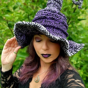 May include: A purple and black witch hat with a curled brim and a tall, pointed crown. The hat is crocheted and has a white and black trim. The hat is worn by a person with purple hair and a crescent moon necklace.