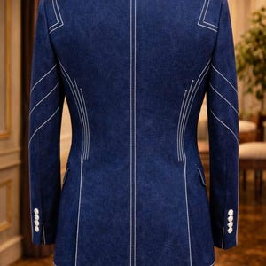 May include: A tailored navy blue blazer with white stitching details. The back of the jacket features vertical and angled white stitching, with white buttons on the sleeves. The blazer is displayed on a mannequin.