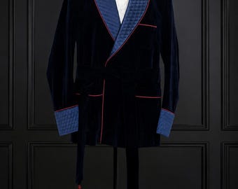 May include: A navy blue velvet smoking jacket featuring a quilted blue shawl collar and matching quilted cuffs. The jacket includes thin red piping along the lapels and pockets, a matching waist tie with red tassels, and a white shirt with a black bow tie.