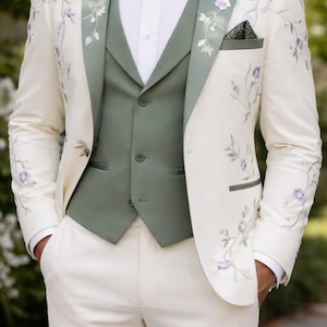 May include: Ivory suit with floral embroidery, featuring a sage green vest and lapel trim. The suit includes matching ivory pants and a black bow tie. A pocket square adds a patterned detail to the ensemble.