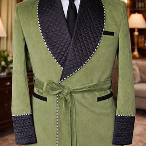 Puede incluir: Bata de terciopelo verde oliva con solapas, puños y ribetes de bolsillo acolchados en negro. La bata presenta un cinturón a juego y pespuntes blancos. Se ve una corbata negra y una camisa blanca.