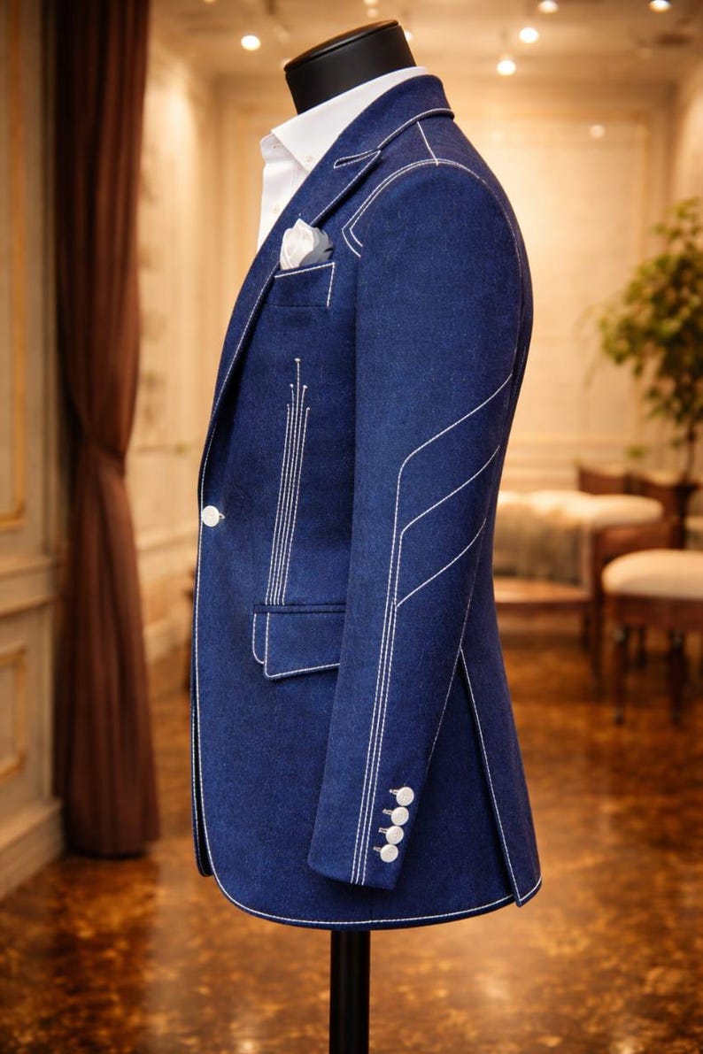May include: A navy blue blazer with white stitching details. The blazer features a white button closure, a pocket square, and white stitching along the seams and cuffs. The jacket is displayed on a mannequin.