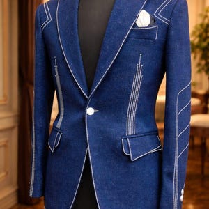 Puede incluir: Un blazer azul oscuro con costuras blancas. El blazer presenta una solapa con muescas, un cierre de un solo botón y un pañuelo de bolsillo. Las costuras blancas delinean los bordes y añaden un elemento decorativo. El blazer se exhibe en un maniquí negro.