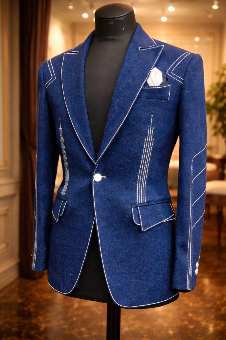 May include: A tailored, dark blue blazer with white stitching accents. The blazer features a notched lapel, a single-button closure, and a pocket square. The white stitching outlines the edges and adds a decorative element. The blazer is displayed on a black mannequin.