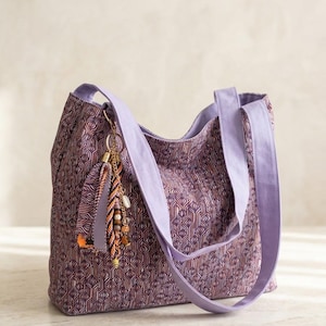 May include: A purple tote bag with a geometric pattern in shades of purple and orange. The bag has lavender straps and a decorative tassel with beads. The bag is designed for carrying items.
