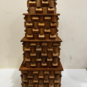 May include: A stack of four square wooden boxes with a medium brown finish. Each box features a repeating geometric pattern of protruding, faceted blocks on its sides. The boxes are arranged in a vertical tower, decreasing in size from bottom to top.