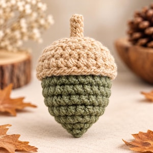 Può includere: Un ornamento a forma di ghianda realizzato all'uncinetto, caratterizzato da una cupola beige testurizzata con un piccolo stelo e una base a coste color verde oliva. La lavorazione presenta un motivo a maglia uniforme e arrotondato.