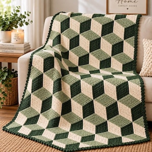 May include: A rectangular crochet blanket featuring a geometric 3D cube pattern in shades of dark forest green, sage green, and cream. The blanket has a scalloped border edge and is draped over a sofa, displaying a repeating optical illusion design.