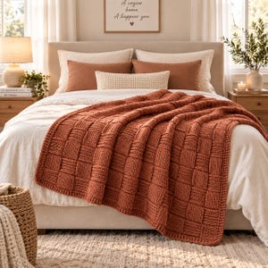 Puede incluir: Una manta de punto color óxido con un patrón de tejido de cesta texturizado sobre una cama. La cama cuenta con un edredón blanco, dos almohadas blancas grandes, dos almohadas rectangulares color óxido y un pequeño cojín lumbar color crema.