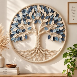 May include: A circular wall hanging featuring a macrame tree of life design. The tree has a braided trunk and roots made of beige cord, with numerous individual leaves in shades of cream, light blue, and dark blue attached to the branches.