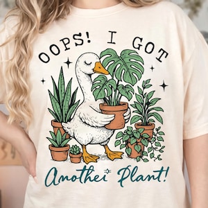 Könnte beinhalten: Cremefarbenes T-Shirt mit einer Illustration einer weißen Gans, die eine Topfpflanze trägt. Der Text "OOPS! I GOT Another Plant!" ist oberhalb und unterhalb der Gans in schwarzer und türkisfarbener Schrift gedruckt. Verschiedene Topfpflanzen umgeben die Gans.