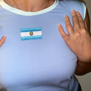 May include: A light blue short-sleeved top featuring a round neckline with a pale mint green trim. Centered on the chest is a rectangular embroidered patch displaying the national flag of Argentina, characterised by horizontal light blue and white stripes with a central yellow sun emblem.