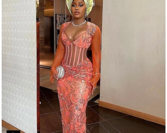 May include: A floor-length coral orange gown featuring a corset-style bodice with sheer panels, long sheer sleeves, and intricate sequin embroidery. The ensemble includes a matching structured headpiece and a small silver clutch accessory.