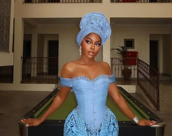 May include: A light blue off-the-shoulder corset dress featuring a sweetheart neckline with silver embellishments, a front button closure, and intricate sequin patterns. The ensemble includes a matching light blue headwrap with a pleated design.