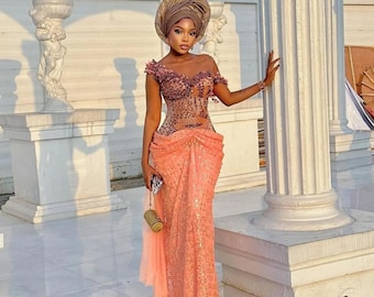 May include: A floor-length peach-colored gown featuring a sheer, embellished bodice with dark floral accents and an off-the-shoulder neckline. The skirt is made of textured lace fabric with a draped waist detail. A matching headwrap and small gold clutch.