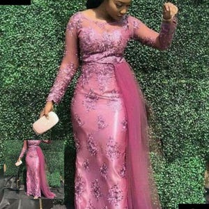 Puede incluir: Un vestido largo y rosa formal con intrincados bordados florales y mangas transparentes. Una tela transparente a juego cae de la cintura. El vestido se combina con un clutch blanco y un pañuelo verde.